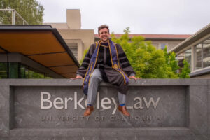 Javier Mabrey sitting on a stone wall that contains text. "Berkeley Law University of California"